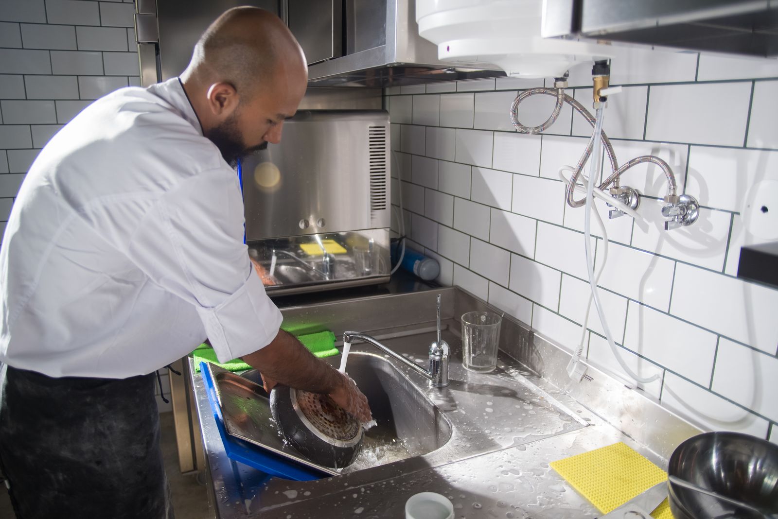 A chef washing cookware at a stainless-steel sink in a tiled commercial kitchen