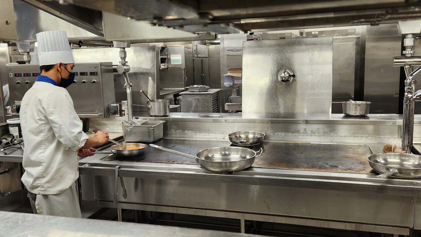 A chef in a face mask and toque cooking at a stainless-steel line in a commercial kitchen